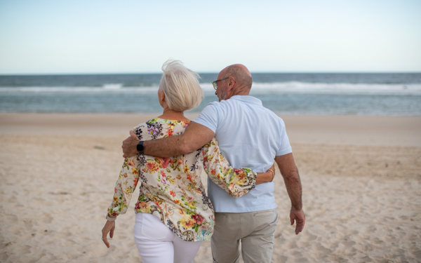 Older couple walking on beach with arms around each other | Icon Cancer Centre